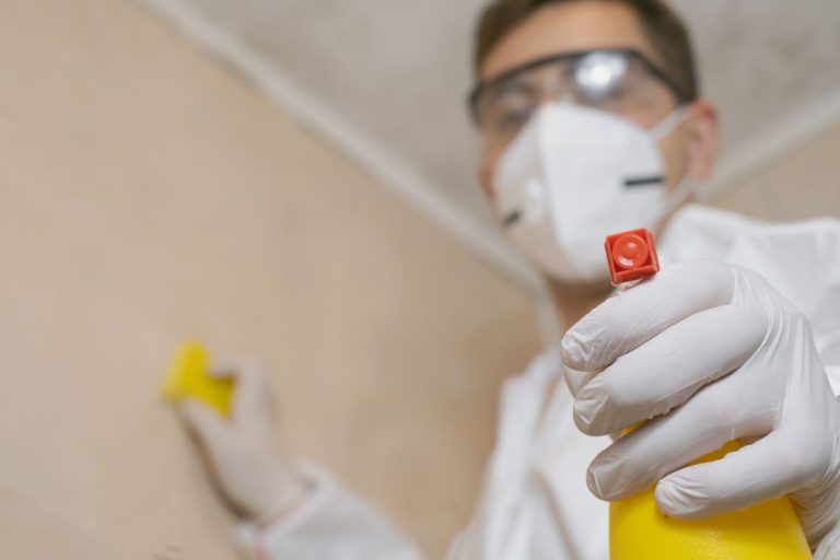 A man dressed in a white coat and gloves is ready with a yellow spray bottle for mold remediation efforts.