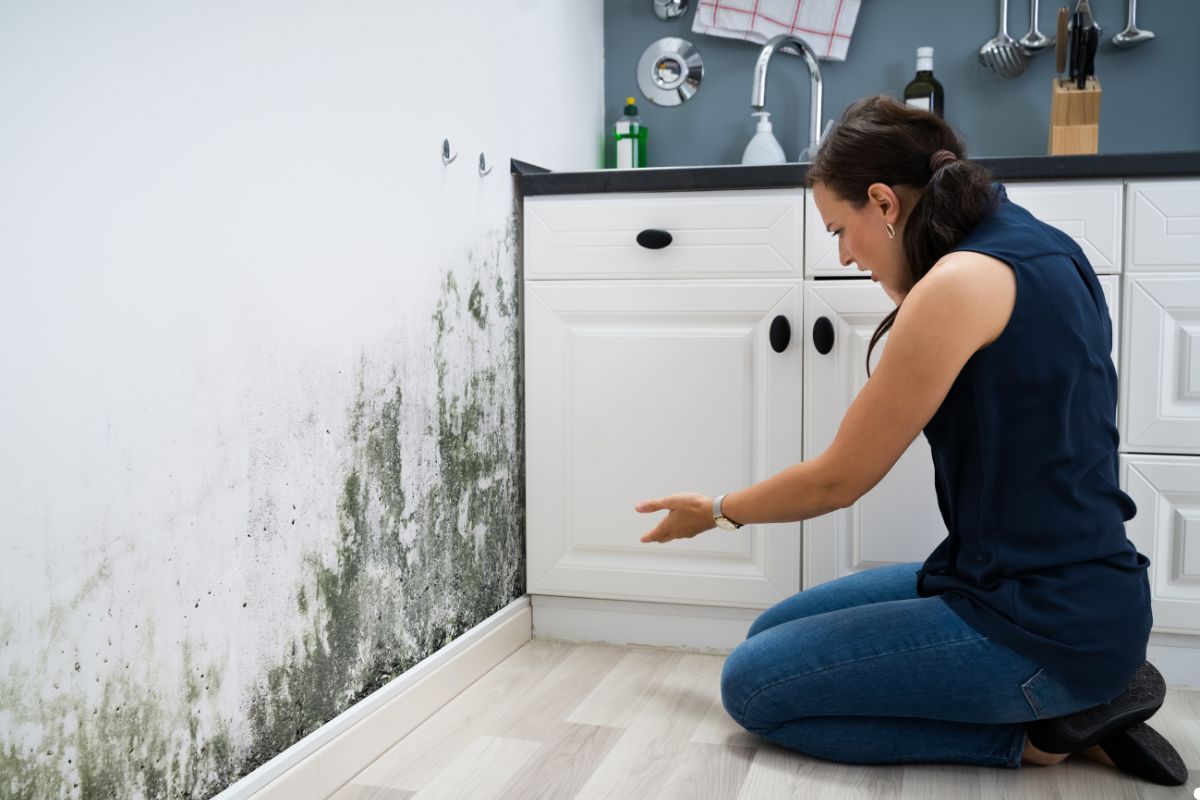 A woman kneels in a kitchen, examining mold on the wall, highlighting a potential health concern.